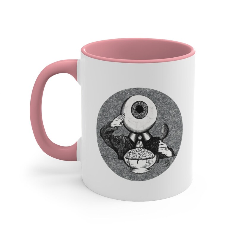 May include: White ceramic coffee mug with a pink rim. The mug features a black and white illustration of an eye with a body, wearing a suit and saluting, eating a bowl of brains.
