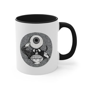 May include: White ceramic coffee mug with a black rim and handle. The mug features a black and white illustration of a person with an eye for a head eating cereal.