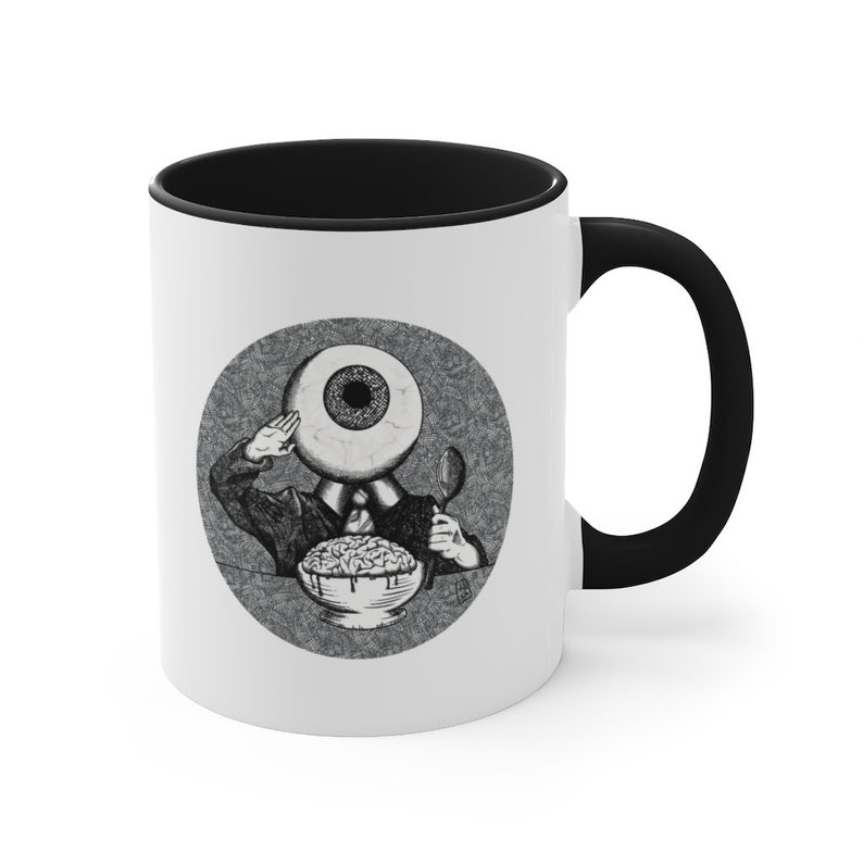 May include: White ceramic coffee mug with a black rim and handle. The mug features a black and white illustration of a person with an eye for a head eating cereal.