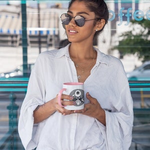 May include: A woman wearing sunglasses and a white button-down shirt holds a pink and white coffee mug with a black and white illustration of an eye on it.
