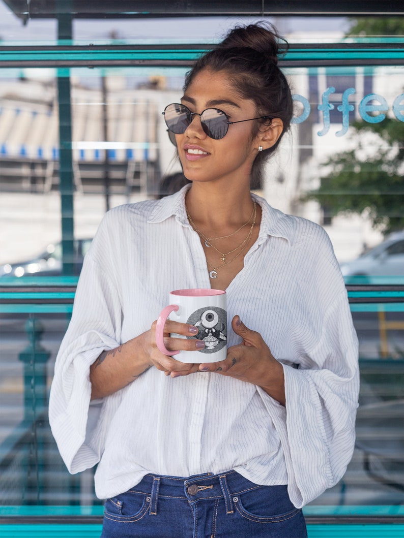 May include: A woman wearing sunglasses and a white button-down shirt holds a pink and white coffee mug with a black and white illustration of an eye on it.