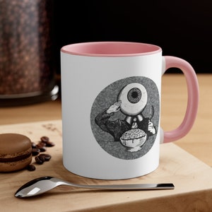 May include: A white ceramic coffee mug with a pink rim and handle. The mug features a black and white illustration of a person with an eye for a head, wearing a suit and eating cereal.