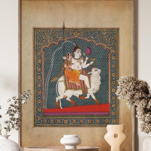 May include: A framed artwork depicting a Hindu deity riding a white bull. The artwork is set against a blue background with intricate gold detailing. The artwork is displayed on a wooden shelf with other decorative items.