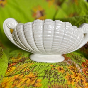 May include: A white ceramic bowl with a shell-like design and two handles. The bowl is sitting on a green and brown patterned fabric.
