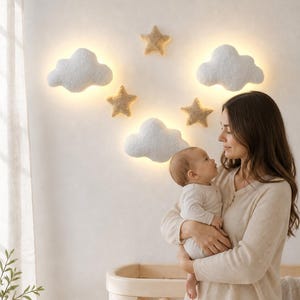 Może przedstawiać: Zestaw lamp ściennych składający się z trzech białych lamp w kształcie chmur oraz trzech beżowych dekoracji w kształcie gwiazd. Lampy chmury emitują miękkie, ciepłe światło zza swoich powierzchni, rozmieszczone wraz z gwiazdami na ścianie.