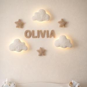 May include: A nursery scene featuring a white changing table with a plush changing pad. Above, illuminated cloud and star wall decor surrounds the name "OLIVIA" in soft, textured letters. A basket with a teddy bear is below.