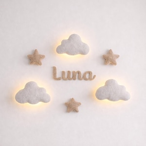 May include: A nursery wall decoration featuring the name "Luna" in light brown letters, surrounded by felt clouds and stars. The clouds have a soft glow. A white crib is visible at the bottom.
