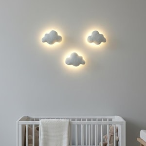 Może przedstawiać: Pokój dziecięcy z białym łóżeczkiem, szarym materacem, białym kocem i misiem. Nad łóżeczkiem trzy szare lampy ścienne w kształcie chmur są podświetlone na szarej ścianie.