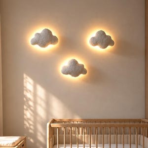Op de afbeelding: Drie witte, wolkvormige wandlampen gemonteerd aan een muur boven een houten wieg. Elke lamp heeft een getextureerd oppervlak en straalt een warme, zachte gloed uit van achter de randen, wat een halo-effect creëert op het muuroppervlak.