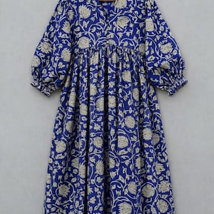 Op de afbeelding: Een midi-jurk in diepblauw met een herhalend beige bloemenpatroon. Het kledingstuk is voorzien van een rucheskraag, een knoopsluiting aan de voorzijde en driekwart pofmouwen met elastische boorden, hangend aan een houten kledinghanger.