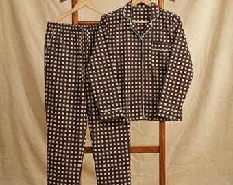 Könnte beinhalten: Ein braun-weiß kariertes Pyjama-Set, bestehend aus einem Hemd mit Knöpfen und einer Hose, hängt an einem hölzernen Kleiderständer. Der Pyjama hat weiße Paspeln und ist aus einem weichen Stoff gefertigt. Der Hintergrund ist ein neutrales Beige.