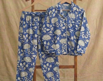 Könnte beinhalten: Blaues Pyjama-Set mit floralem Muster, bestehend aus einem Hemd mit Knöpfen und einer Hose mit Kordelzug, hängt an einem Holzkleiderbügel. Das florale Design ist hellbeige, mit weißer Paspelierung.
