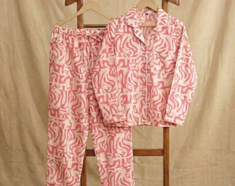 May include: A two-piece pajama set featuring a long-sleeved button-down shirt and matching trousers. The garment is made of a light-colored fabric with an all-over abstract pattern in a vibrant pink hue. The set is displayed on a wooden hanger.