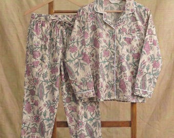 Könnte beinhalten: Beige Pyjama-Set mit rosa und grünen Blumenmustern. Das Set besteht aus einem Hemd mit Knöpfen und einer Hose mit Kordelzug, beide mit weißem Paspel versehen. Der Pyjama hängt an einem hölzernen Kleiderständer.