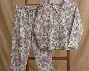 Könnte beinhalten: Ein zweiteiliges Pyjama-Set mit floralem Muster. Das Set besteht aus einem Hemd mit Knöpfen und einer Hose mit Kordelzug. Die Pyjamas sind cremefarben mit einem sich wiederholenden Muster aus lila Blumen und grünen Blättern.