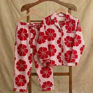May include: A two-piece pajama set featuring a red floral pattern on a light pink striped background. The set includes a button-down shirt with a chest pocket and drawstring pants. The pajamas are hanging on a wooden rack.