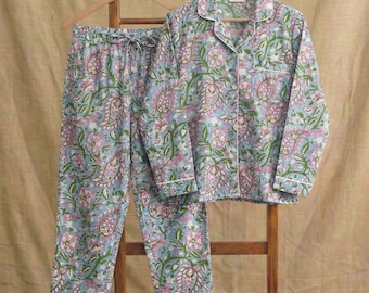 May include: A light blue pajama set featuring a long-sleeved button-down shirt and matching pants. The fabric is decorated with a repeating floral and paisley pattern in shades of pink, green, and white, finished with white piping trim.