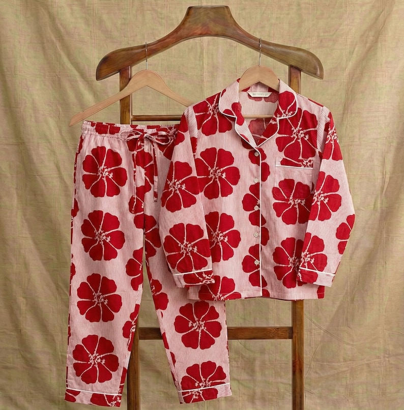 May include: A two-piece pajama set featuring a red floral pattern on a light pink striped background. The set includes a button-down shirt with a chest pocket and drawstring pants. The pajamas are hanging on a wooden rack.