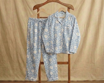 May include: A light blue pajama set featuring a white floral pattern. The set includes a long-sleeved button-down shirt with a collar and a chest pocket, paired with matching loose-fitting trousers. The garments are displayed on a wooden hanger.