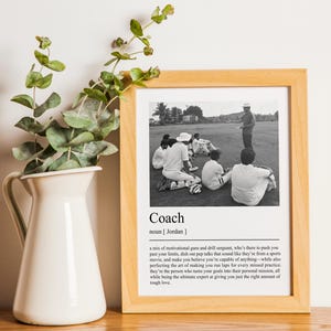 May include: A black and white photo of a coach giving instructions to a group of athletes. The photo is framed in a light wood frame and is accompanied by a definition of the word "coach" in a black and white font.