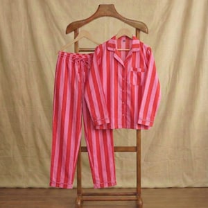May include: A striped pyjama set in red and pink. The set comprises a button-down shirt with a chest pocket and drawstring trousers. The pyjamas are displayed on a wooden hanger and stand against a neutral backdrop.