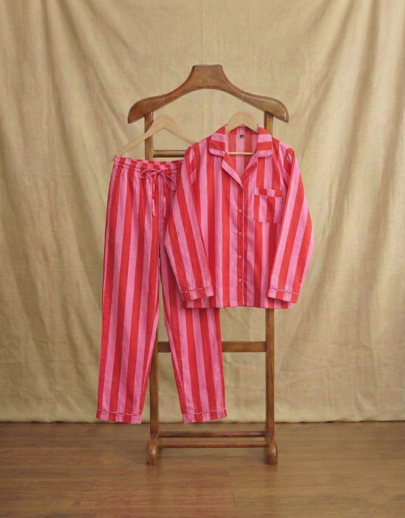 May include: A striped pyjama set in red and pink. The set comprises a button-down shirt with a chest pocket and drawstring trousers. The pyjamas are displayed on a wooden hanger and stand against a neutral backdrop.