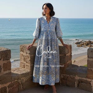 May include: A light blue floral print dress with long sleeves and a high neckline. The dress features a flowing skirt and is worn by a person standing on a stone wall overlooking the ocean. The text "Cadore Print" and "Slow Fashion, True Comfort" are visible.