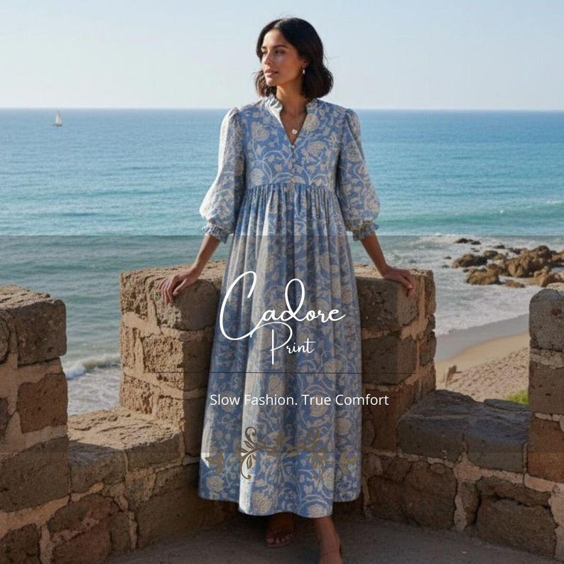 May include: A light blue floral print dress with long sleeves and a high neckline. The dress features a flowing skirt and is worn by a person standing on a stone wall overlooking the ocean. The text "Cadore Print" and "Slow Fashion, True Comfort" are visible.