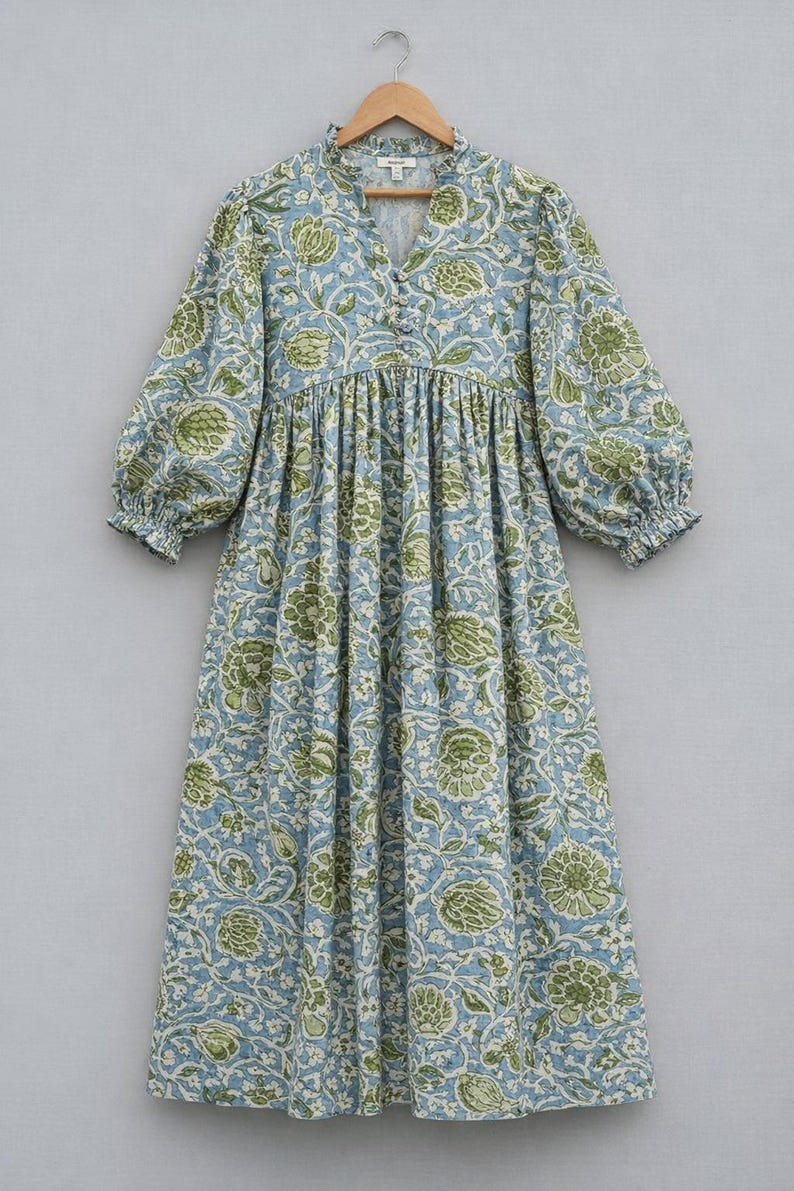 May include: A midi-length dress featuring a light blue base with a repeating green and white floral pattern. The dress has a V-neckline with a small ruffle collar, a button-front placket, and three-quarter length puff sleeves with elastic cuffs.