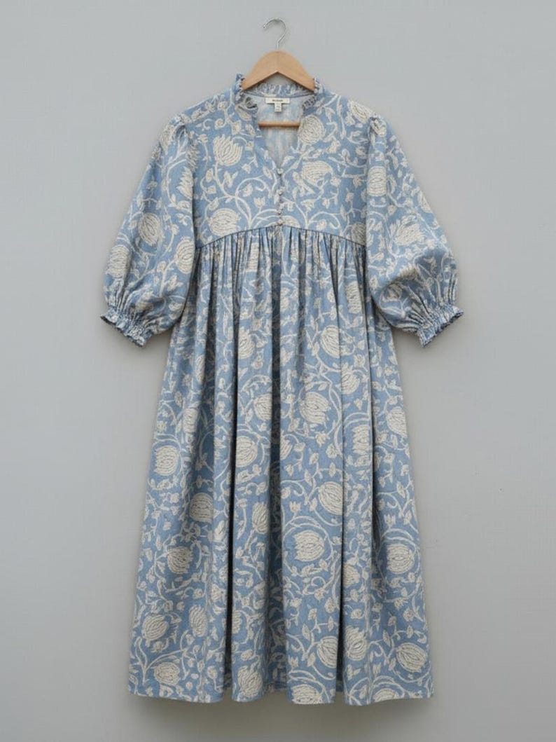 May include: A light blue dress with a white floral pattern. The dress has a high ruffled collar, a V-neck, and three-quarter sleeves with elastic cuffs. The dress is hanging on a wooden hanger.