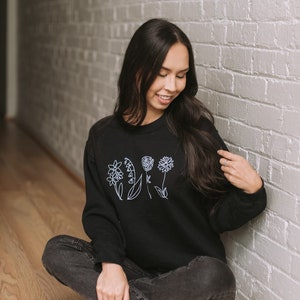 May include: A woman wearing a black sweatshirt with a white outline of flowers on the front. She is sitting on a wooden floor with her legs crossed and wearing black jeans and white sneakers.