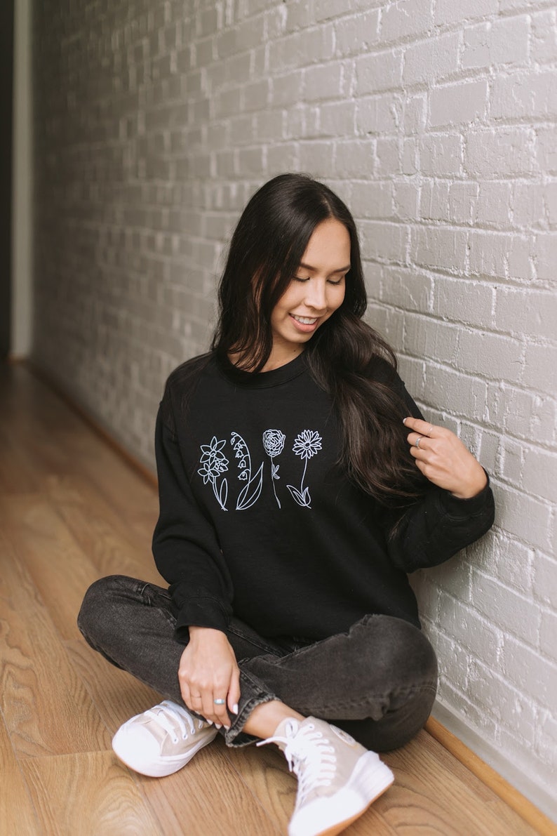 May include: A woman wearing a black sweatshirt with a white outline of flowers on the front. She is sitting on a wooden floor with her legs crossed and wearing black jeans and white sneakers.