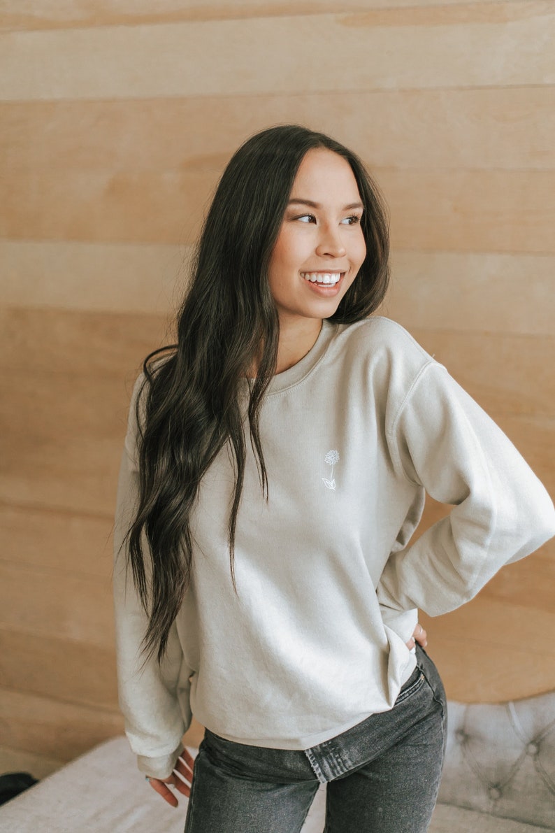 May include: A woman wearing a light gray sweatshirt with a small white flower embroidered on the front. She is wearing dark wash jeans and has long dark hair.