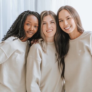 May include: Three women wearing beige sweatshirts are standing together and smiling. The woman in the middle is wearing a sweatshirt with a small flower design on the front.