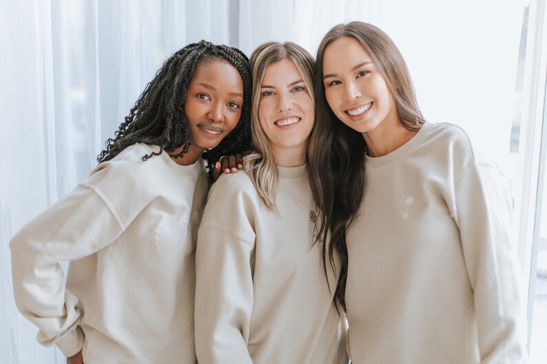 May include: Three women wearing beige sweatshirts are standing together and smiling. The woman in the middle is wearing a sweatshirt with a small flower design on the front.