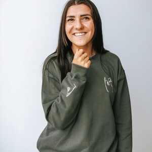 May include: A woman wearing a dark green sweatshirt with a white line drawing of a hand holding a flower on the front. The sweatshirt has a white line drawing of a flower on the left sleeve.