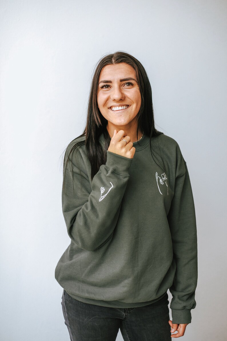 May include: A woman wearing a dark green sweatshirt with a white line drawing of a hand holding a flower on the front. The sweatshirt has a white line drawing of a flower on the left sleeve.