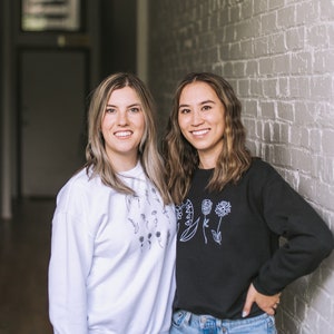 May include: Two women wearing black and white sweatshirts with floral designs. The woman on the left is wearing a white sweatshirt with black floral designs. The woman on the right is wearing a black sweatshirt with white floral designs. Both women are smiling and looking at the camera.