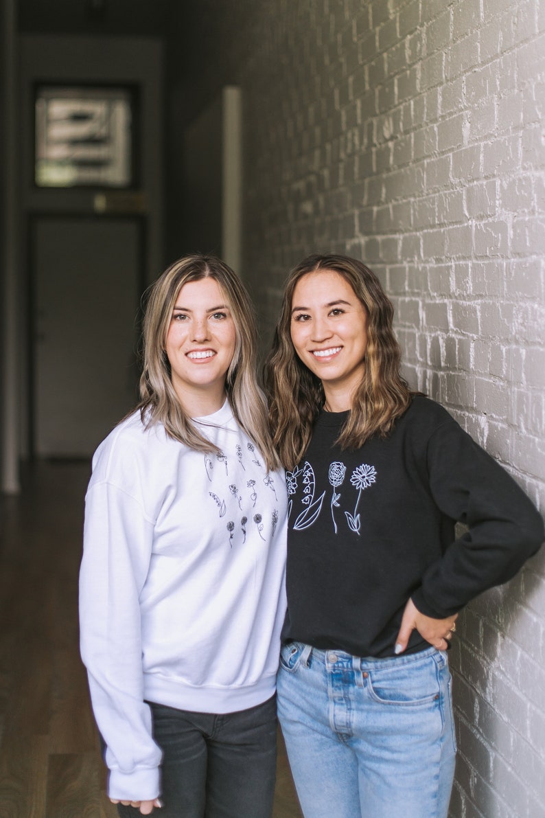 May include: Two women wearing black and white sweatshirts with floral designs. The woman on the left is wearing a white sweatshirt with black floral designs. The woman on the right is wearing a black sweatshirt with white floral designs. Both women are smiling and looking at the camera.
