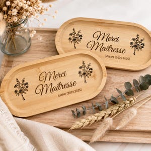 Puede incluir: Dos bandejas ovaladas de bambú con la inscripción "Merci Maîtresse" e ilustraciones florales. Las bandejas están grabadas con nombres y fechas. Se encuentran sobre una superficie de madera.