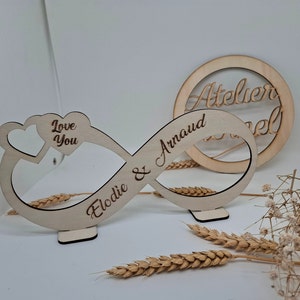 Peut inclure: Un symbole d'infini en bois avec les noms "Elodie & Arnaud" et "Love You" gravés dessus. Un cercle en bois séparé avec le texte "Atelier Novel" est également dans l'image.