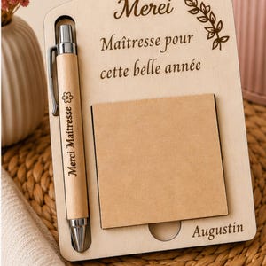 Peut inclure: Un organiseur de bureau en bois avec un stylo, un bloc-notes et du texte gravé. Le stylo est marron clair avec des accents argentés et les mots "Merci Maitresse" sont imprimés sur le stylo. Le bloc-notes est un carré de papier marron clair. Le texte sur l'organisateur dit "Merci Maîtresse pour cette belle année" et "Augustin".