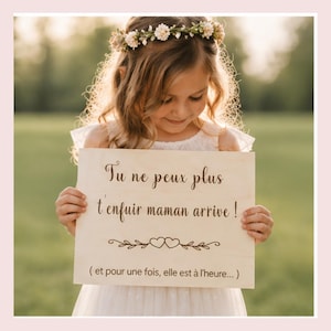 Peut inclure: Une jeune fille portant une couronne de fleurs et une robe blanche tient une pancarte en bois avec du texte en français. La pancarte porte les mots "Tu ne peux plus t'enfuir maman arrive !" et des éléments décoratifs. L'arrière-plan est flou avec la lumière naturelle.