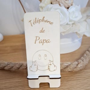 Peut inclure: Support de téléphone en bois clair avec le texte "Téléphone de Papa" et un visage souriant avec des pouces levés. Le support a un design bicolore et une fente pour un téléphone.