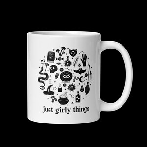 May include: White ceramic mug featuring a black and white illustration of witchy symbols, including a skull, potion bottles, and a witch's hat. The text "just girly things" is printed below the illustration.