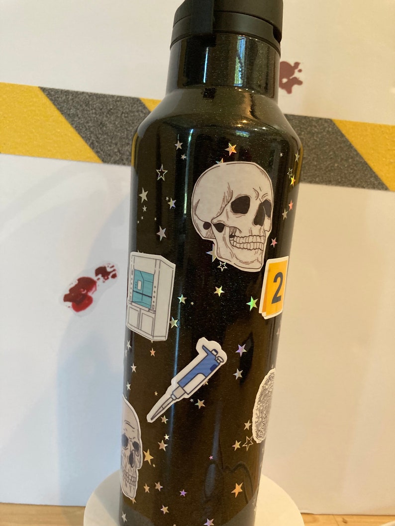 May include: Black stainless steel water bottle with a skull, stars, and other science-themed stickers. The stickers include a pipette, a lab coat, and a number "2".
