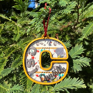 May include: A yellow letter "C" Christmas ornament with a red ribbon. The ornament is filled with miniature crime scene-themed objects and the name "Danielle". The background is a Christmas tree.