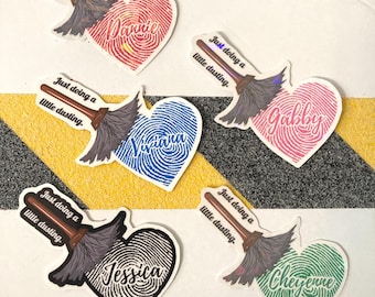 May include: A collection of heart-shaped stickers with fingerprint patterns and personalized names. Each sticker features a broom graphic and the phrase "Just doing a little dusting." The hearts are in various colors, including red, blue, pink, black, and green.