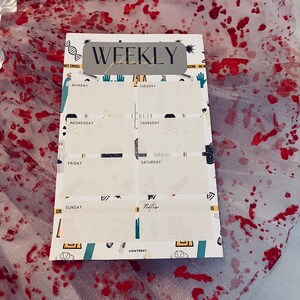 May include: A white weekly planner with a gray "WEEKLY" title bar. The planner has a grid layout with days of the week labeled: Monday, Tuesday, Wednesday, Thursday, Friday, Saturday, and Sunday. The planner has a red, white, and blue color scheme with a blood splatter design.