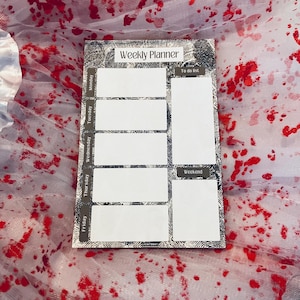 May include: A white weekly planner with a silver glitter background. The planner has spaces for each day of the week, Monday through Friday, and a section for the weekend. The planner also has a "To do list" section. The planner is on a white background with red splatter paint.